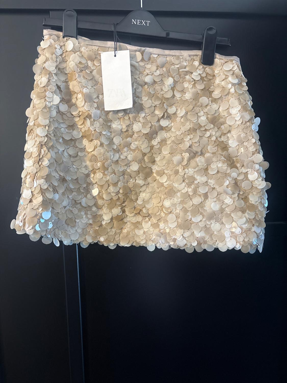 Cream sequinned short skirt on a black hanger with the text NEXT. White label with the text ZARA. Dark background.