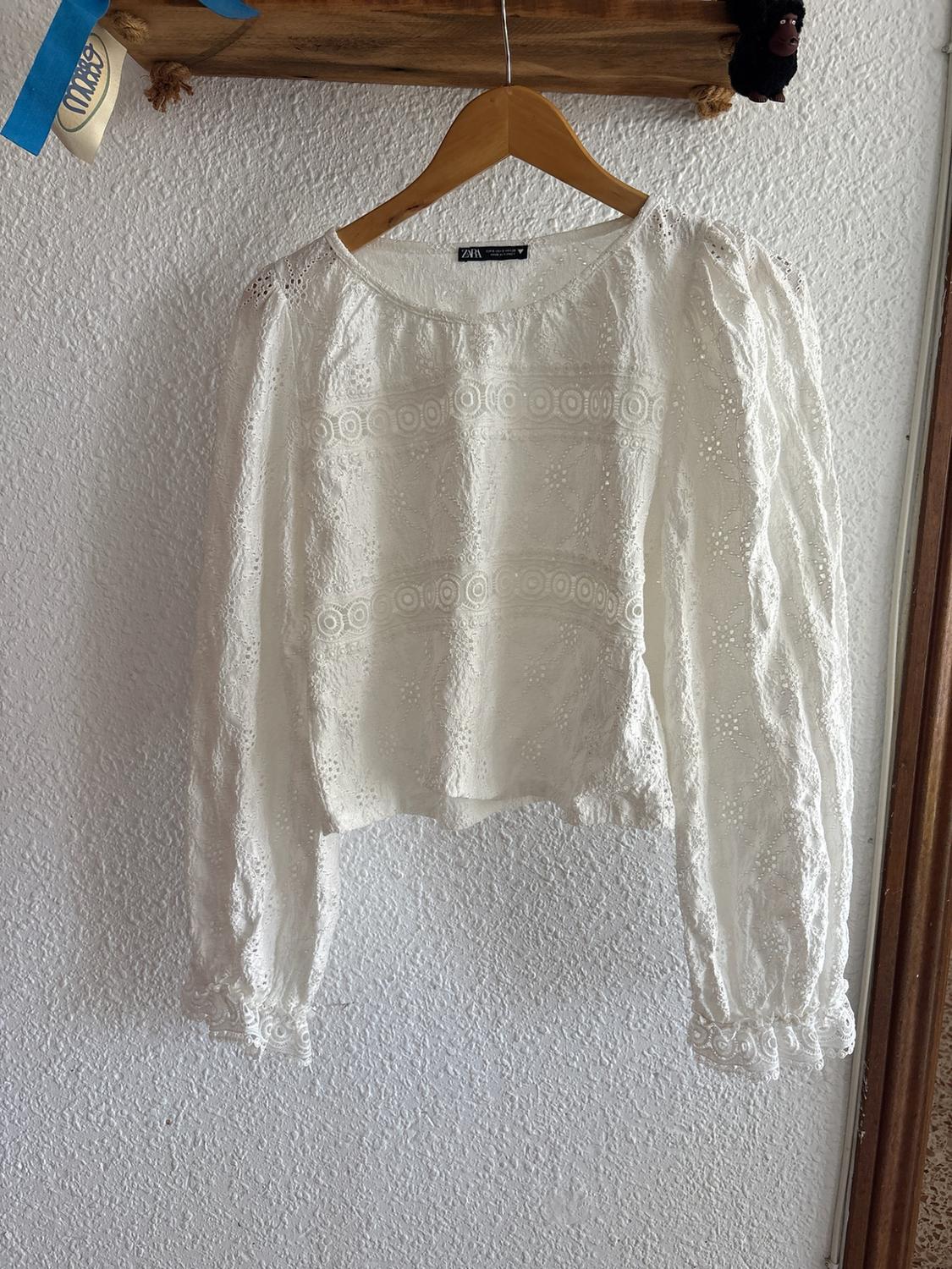White long-sleeved blouse with cut-work embroidery hanging on a wooden hanger against a white wall. Zara label visible.