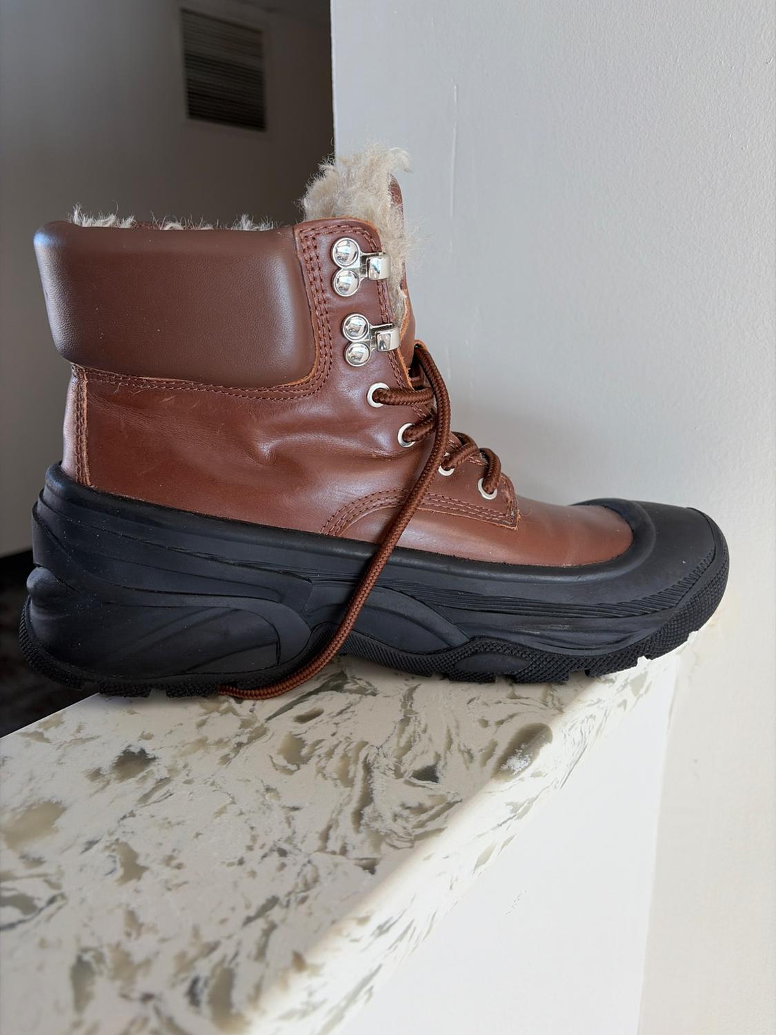 Brown boot with plush lining and black sole on a light marble surface. Brown laces and metallic details.