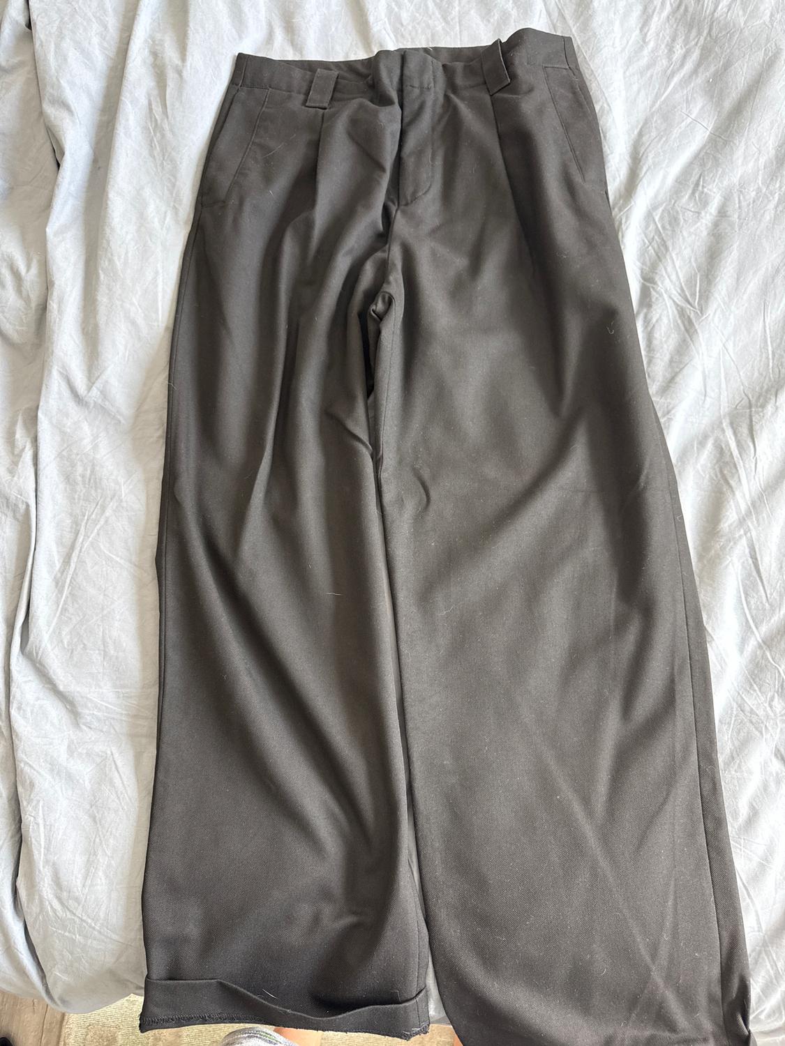 Dark wide-leg trousers on a bed with white sheets. The fabric has a smooth finish and pleats at the top.
