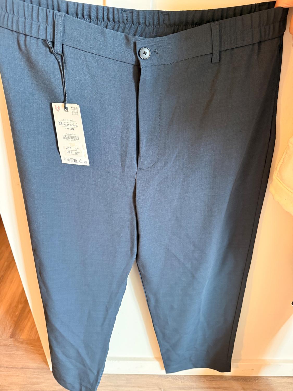 Dark blue pants with an elastic waistband and a hang tag indicating size XL and a price of US$79.99, hanging on a white door.