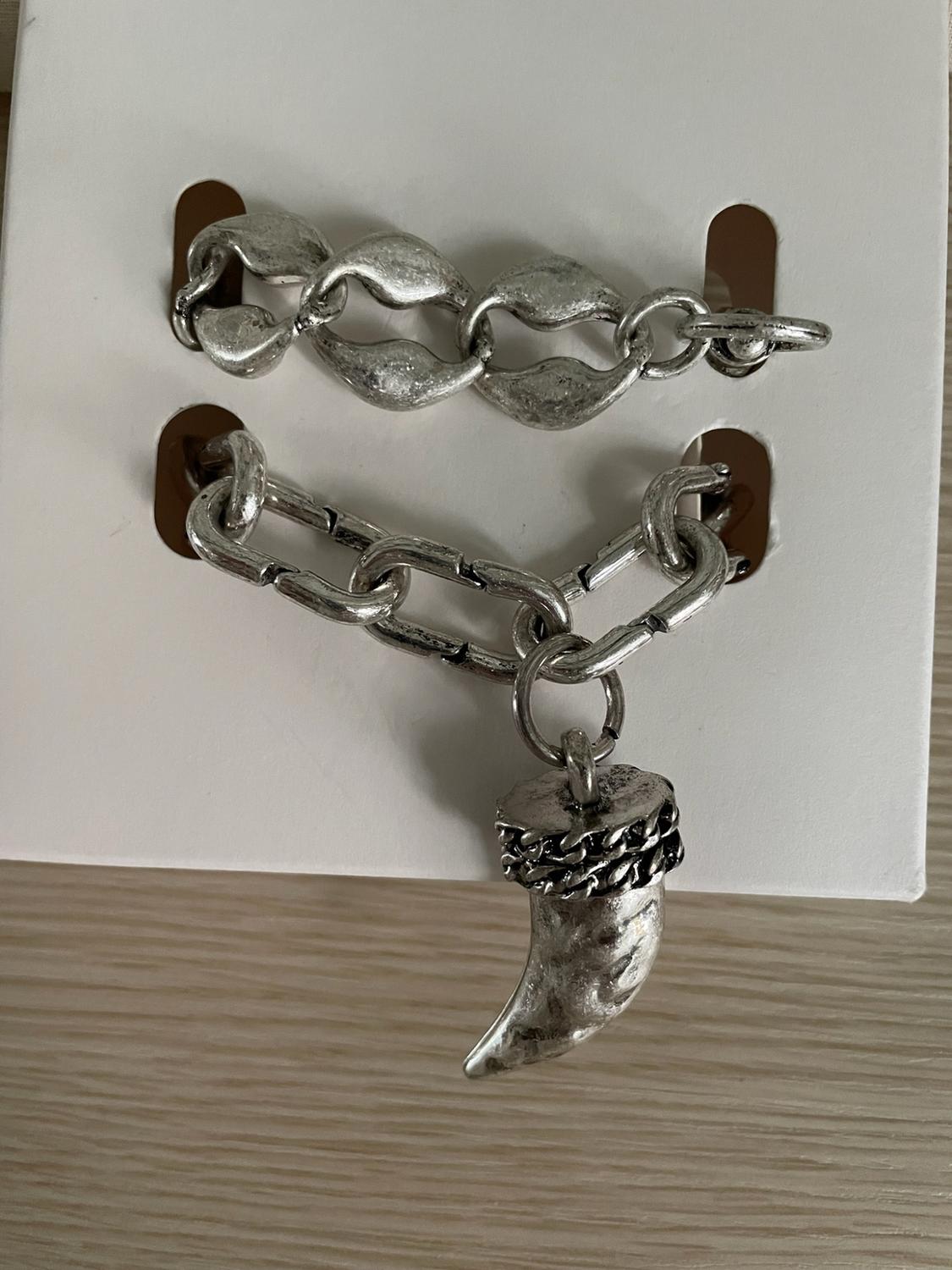 Two silver bracelets on white card. One with chunky links and the other with a textured horn-shaped pendant.