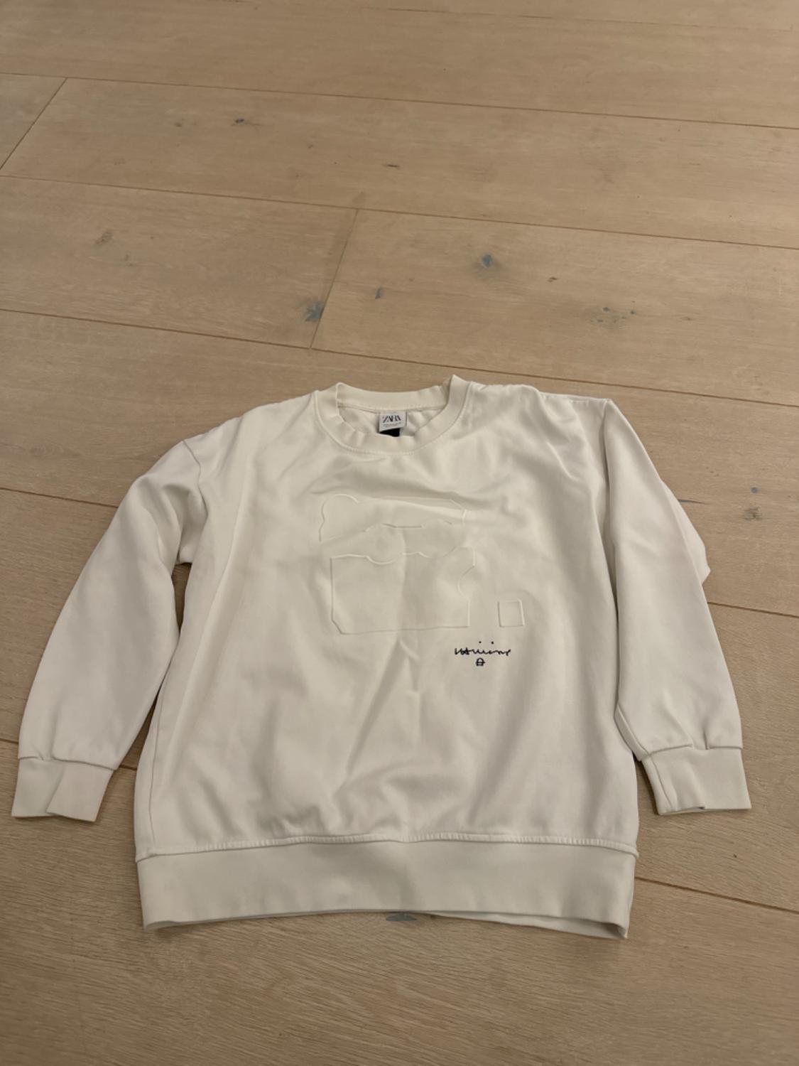 White sweatshirt with embossed design and "Wrinkle" text on the chest, on a light wooden floor.