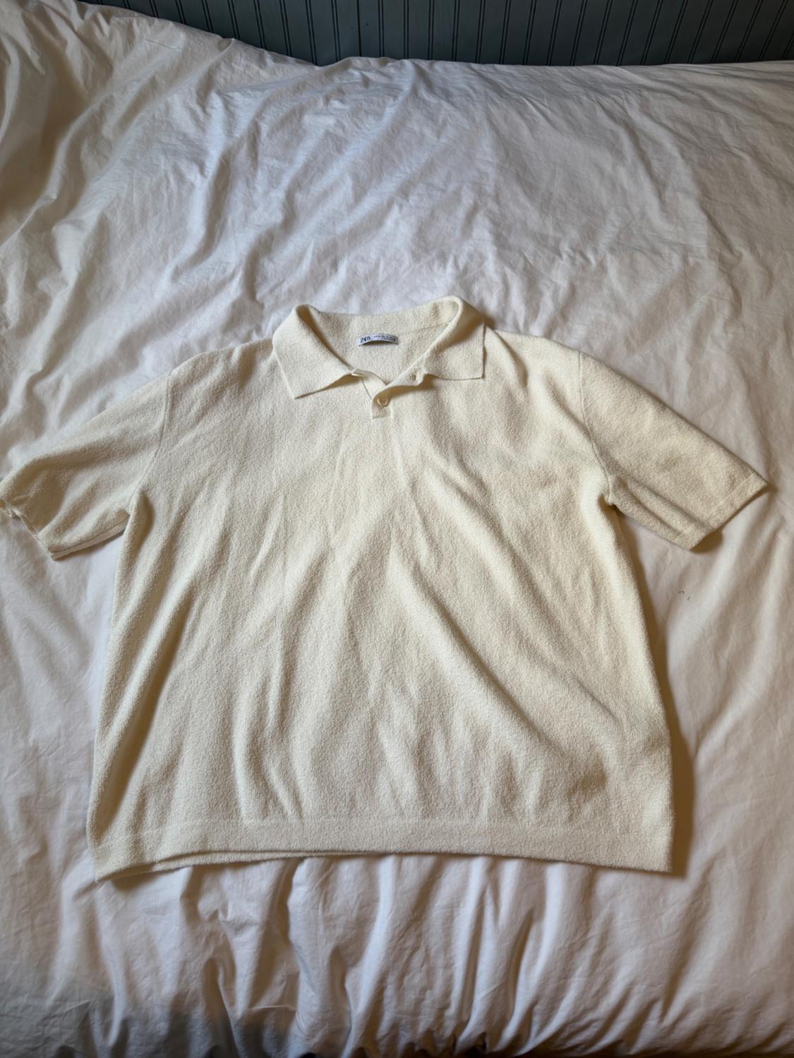 Ecru short-sleeve button-collar polo shirt in textured fabric, resting on wrinkled white sheets.