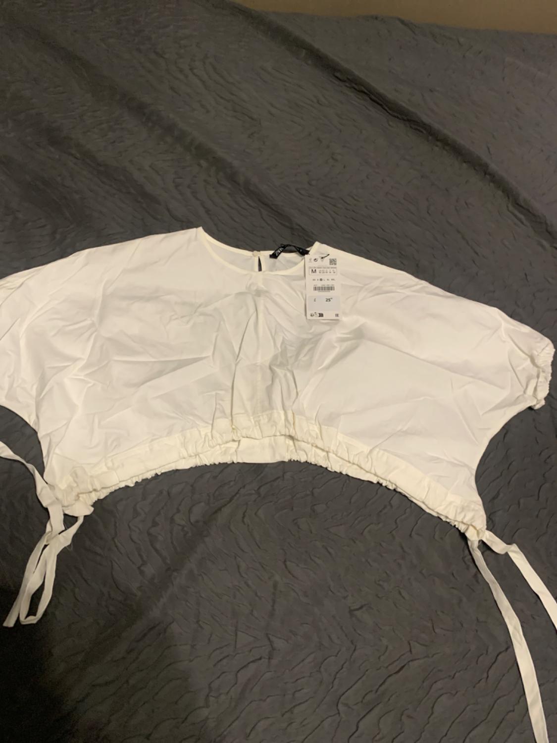 White, short-sleeved blouse with tie at the waist, on a grey bedspread. Visible label with product details.