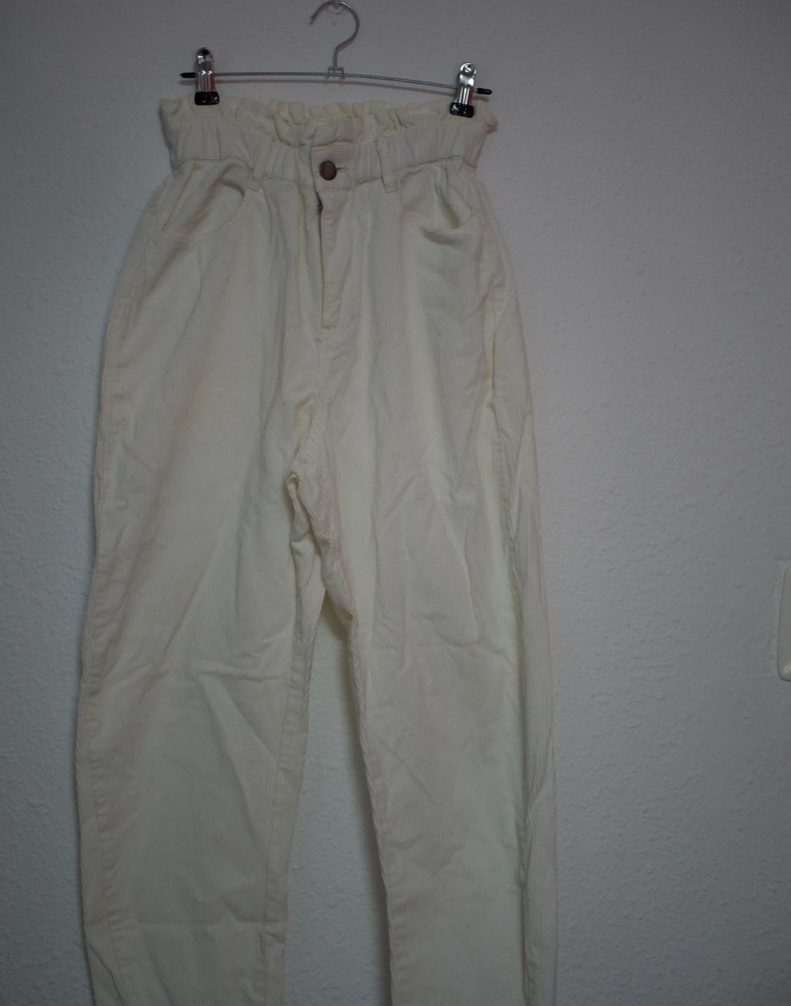 Cream-coloured corduroy trousers with an elasticated gathered waist, hanging on a metal hanger against a white wall.
