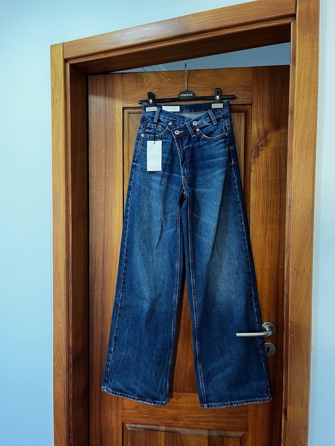 Blue wide-leg jeans with an asymmetrical waist hanging on a wooden door. Hanger with the text FERRACHE.