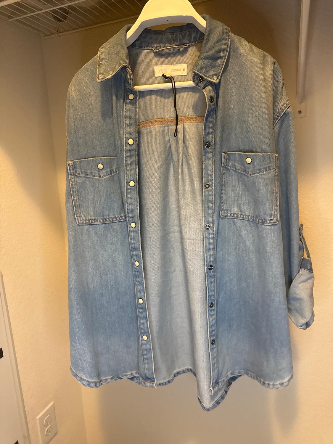 Light blue denim shirt hanging on a white hanger inside a wardrobe. It has front pockets and metal buttons.