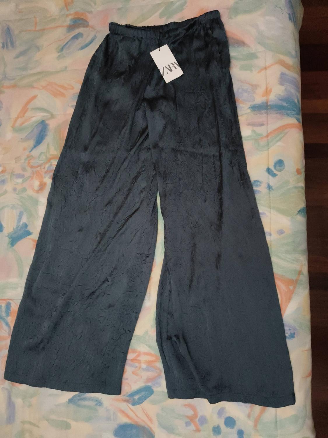 Dark blue wide-leg trousers with a crinkled texture, laid out on a bed with a patterned bedspread. Label with the text ZARA.