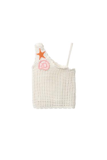 Front view of a white crochet top with starfish appliqués and at symbol on the strap.