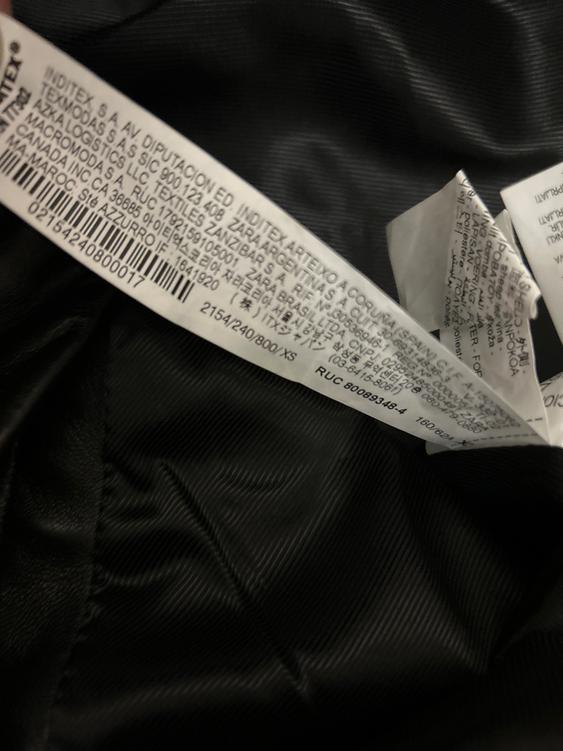 Clothing label with multi-language text on black fabric. Manufacturing details and barcodes are visible.