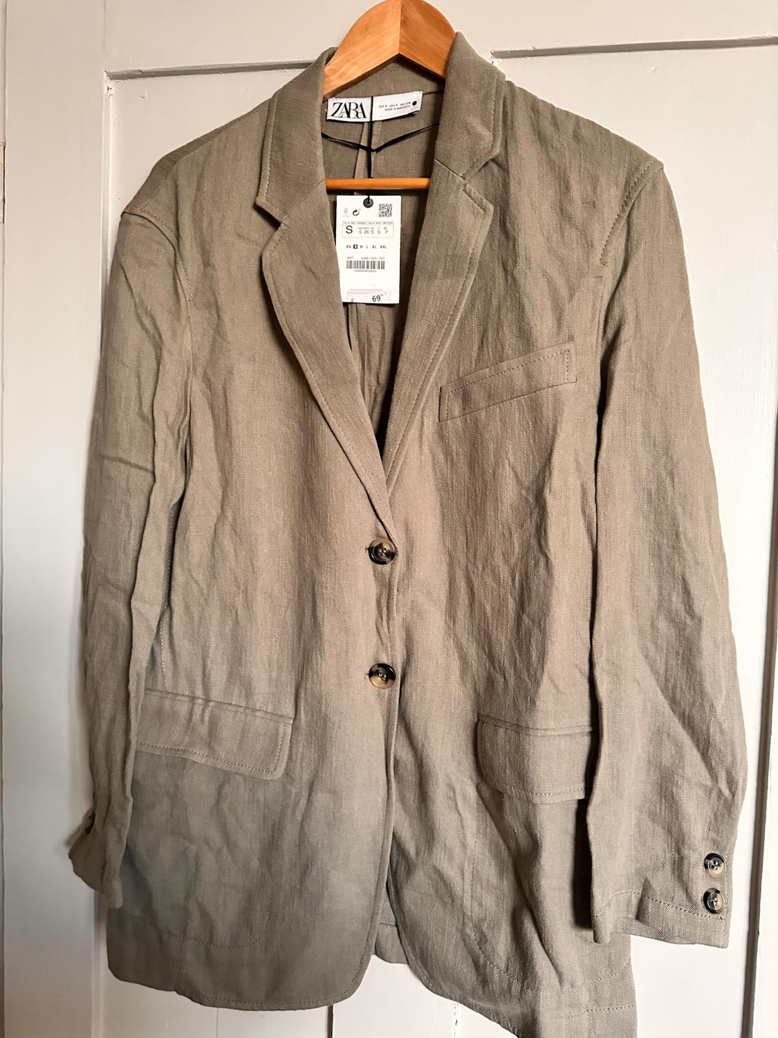 Beige linen jacket hanging on a wooden hanger, with price tag visible. ZARA brand on the neck label.