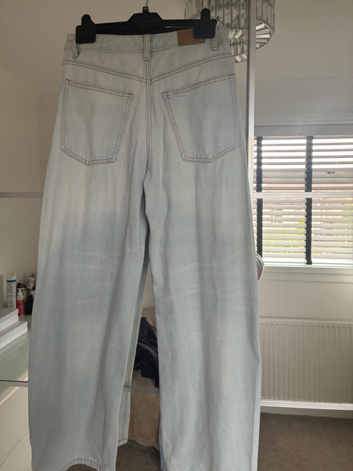 Light-blue denim jeans hanging on a black hanger in front of a mirror. Rear view with two pockets and a brown label.