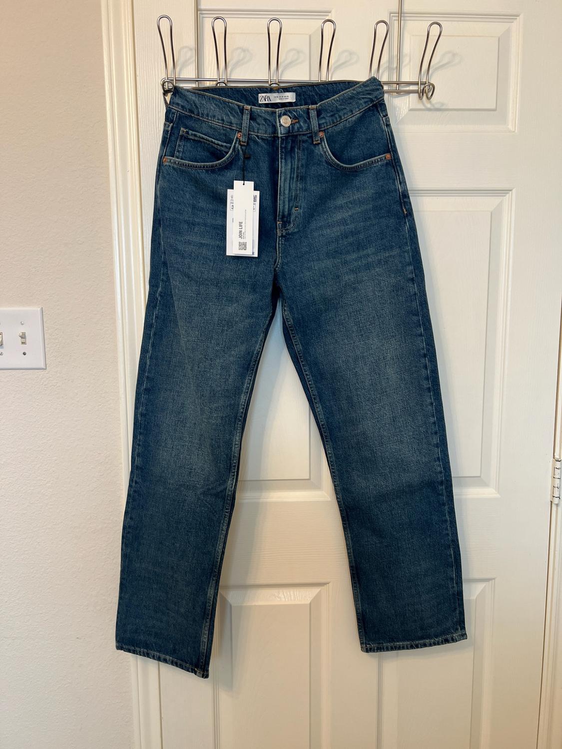 Blue denim trousers hanging from a coat hanger on a white door. Labels bearing the text ZARA and JOIN LIFE can be seen.