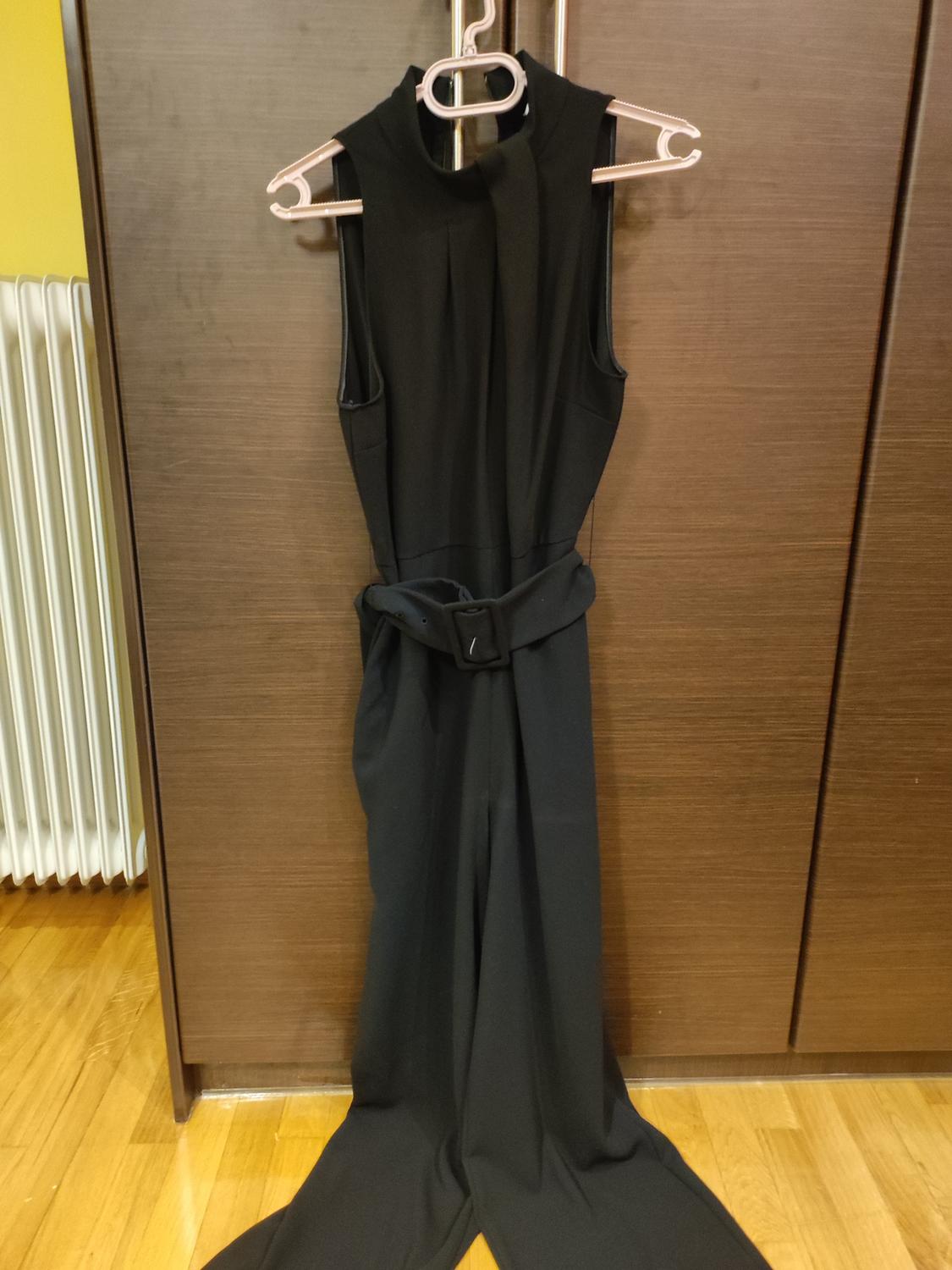 Black sleeveless jumpsuit with belt, hanging on a pink hanger in front of a wooden wardrobe. Wooden floor and radiator in the background.