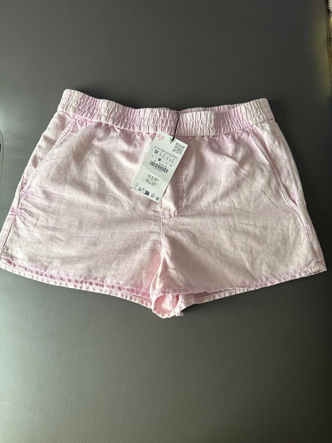 Pink shorts in a soft fabric with an elasticated waistband and side pockets, on a grey surface. Visible label with product details.