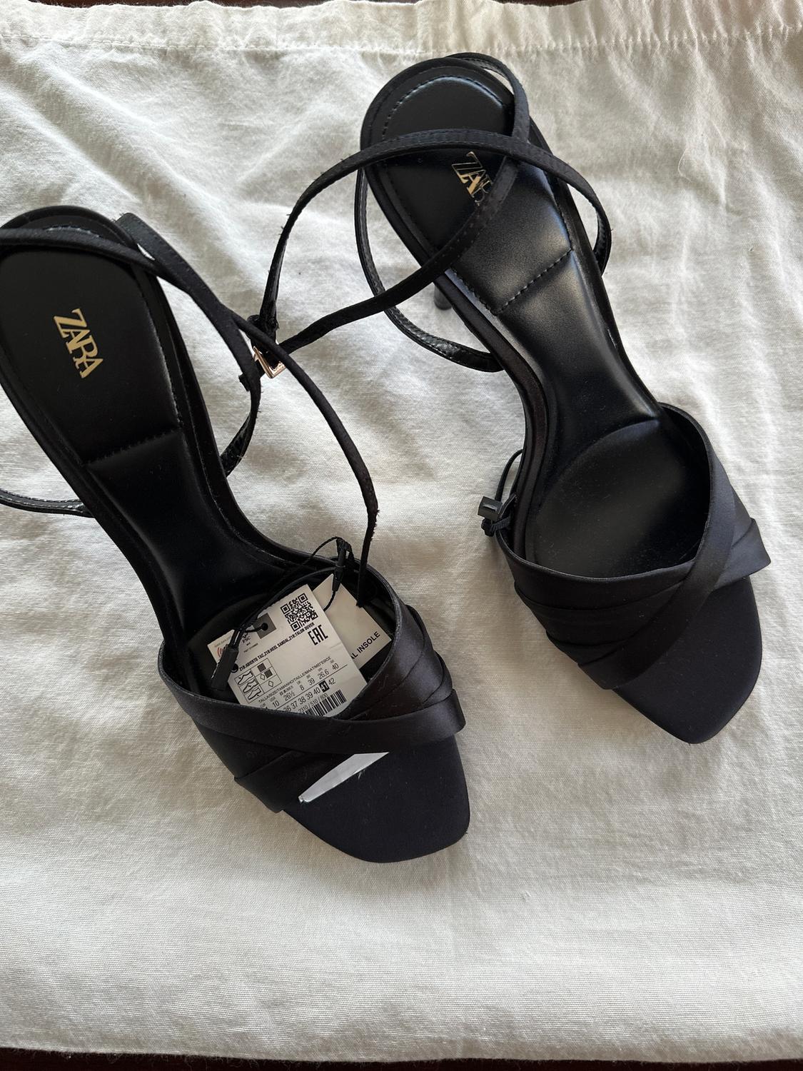 Black high-heeled sandals with crossed straps and a visible label, on white fabric. "ZARA" logo on the insole.