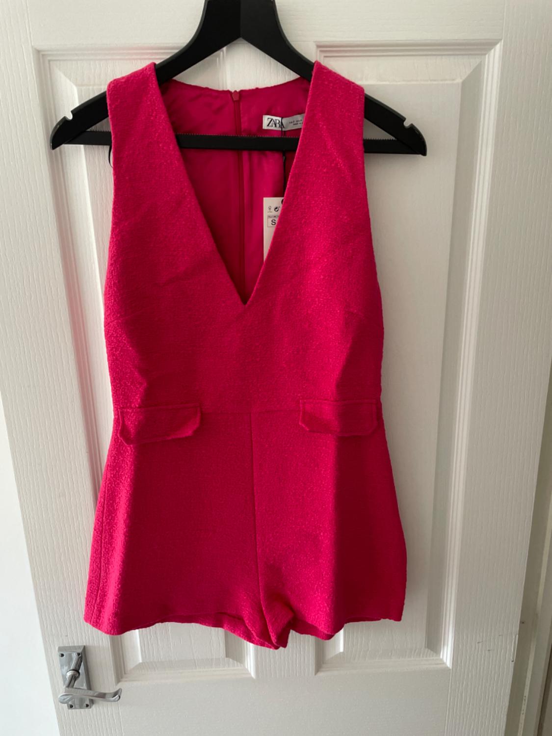 Fuchsia pink playsuit with a V-neck and pockets, hanging on a black hanger on a white door. Labels with the text ZARA and S.