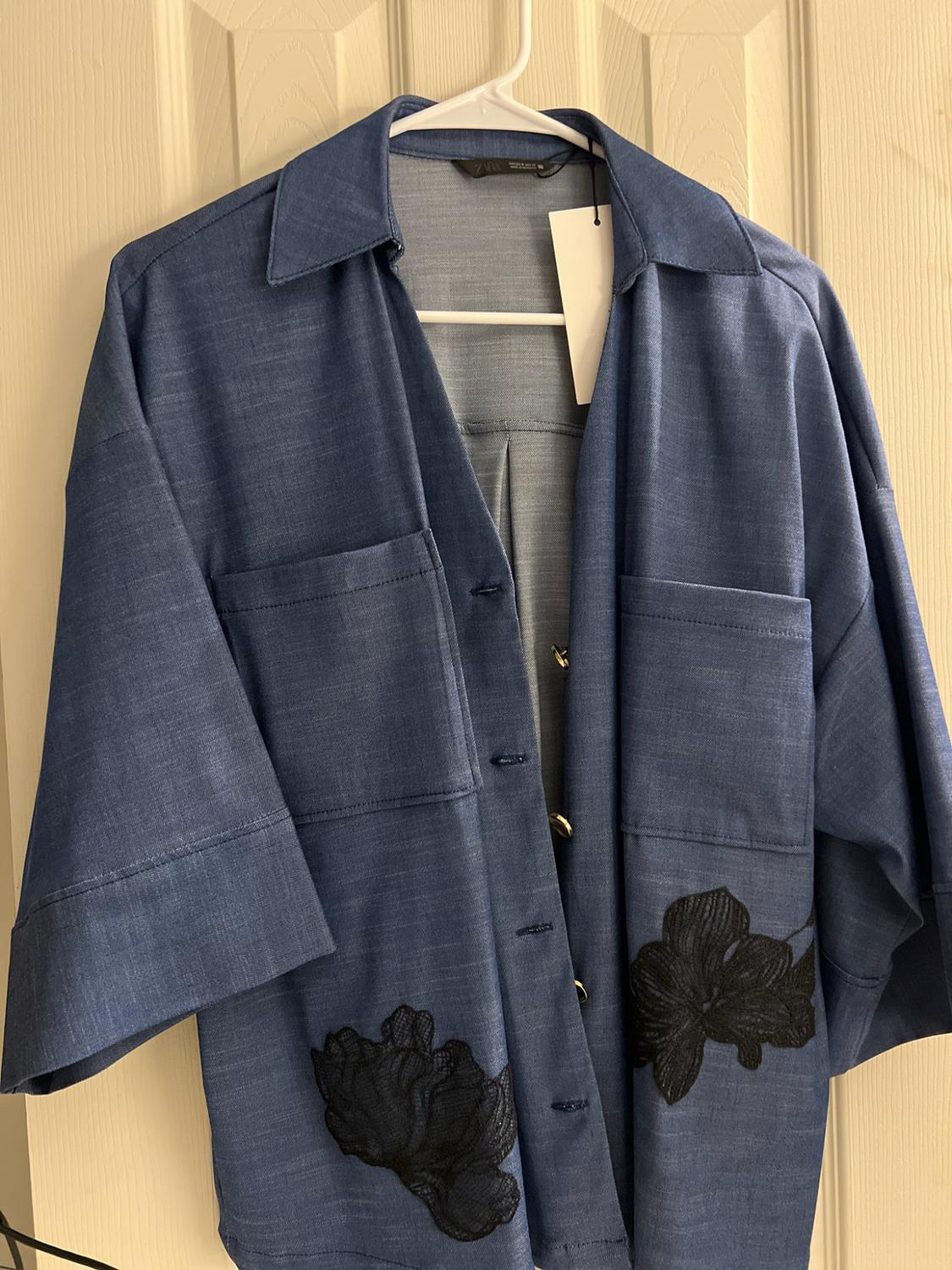 Blue shirt hanging on a white door. It features short sleeves, front pockets, black floral embroidery, and a ZARA label.