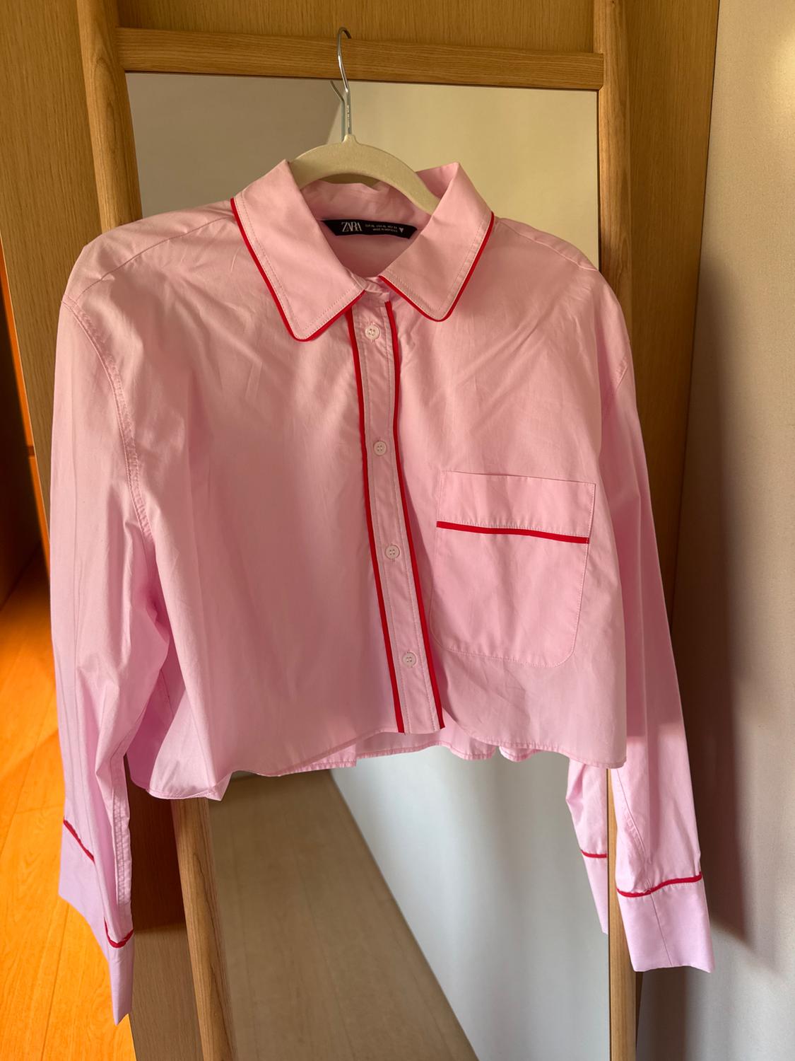 Pink cropped shirt with red trim hanging on a wooden hanger in front of a mirror. Visible label: "Zara".