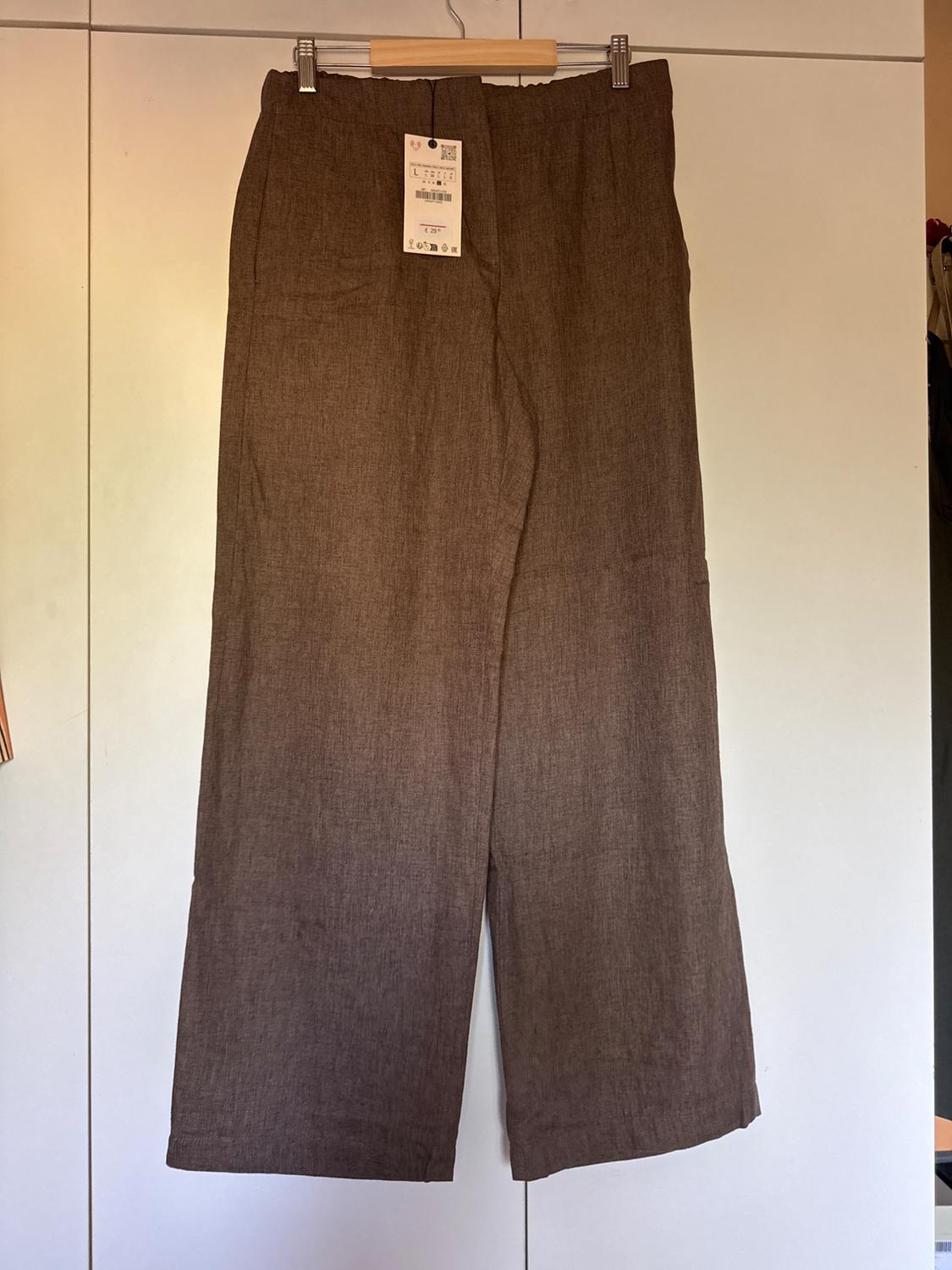 Brown linen trousers hanging on a wooden hanger, with visible label. White wardrobe background.