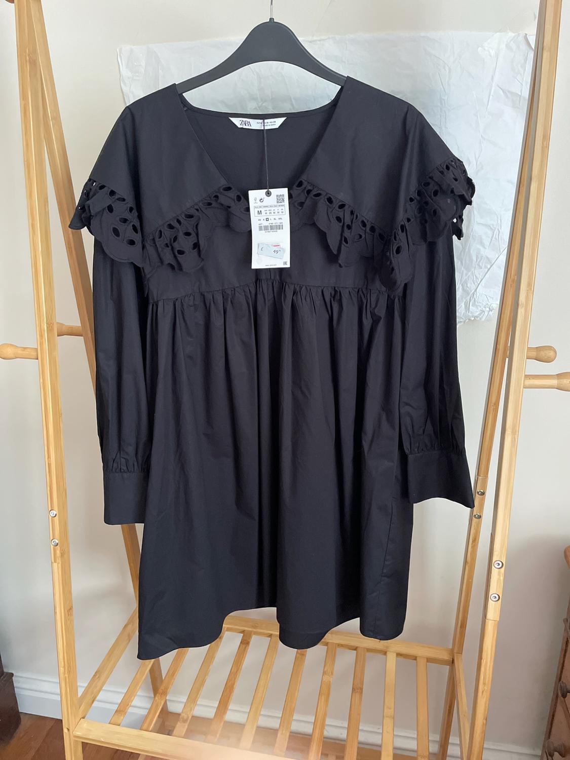 Short black long-sleeved dress with a V-neck and die-cut frill. Hanging on a wooden coat rack with the ZARA label visible.