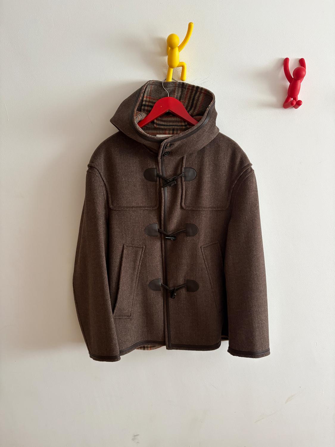 Brown duffle coat with a hood, hanging on a red hanger from a yellow hook on the white wall.