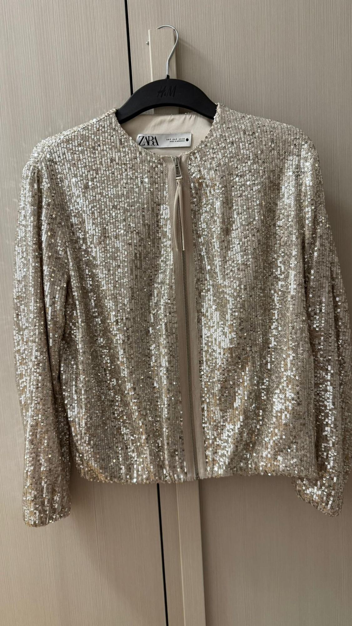 Silver sequin jacket hanging on a black hanger with a visible Zara label, against a light wooden background.