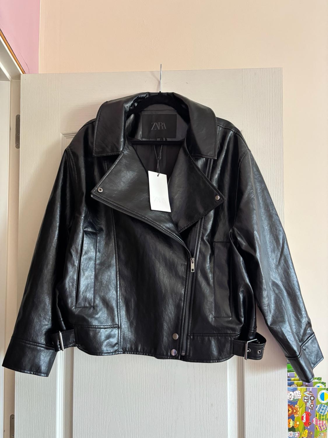 Black jacket with an asymmetrical zip and buckles at the hem, hanging on a white door. Label with the text ZARA visible.