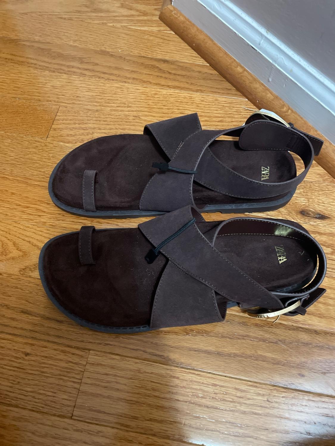 Dark brown suede sandals with crisscross straps, a toe strap, and a gold buckle on a wooden floor. Text zara.