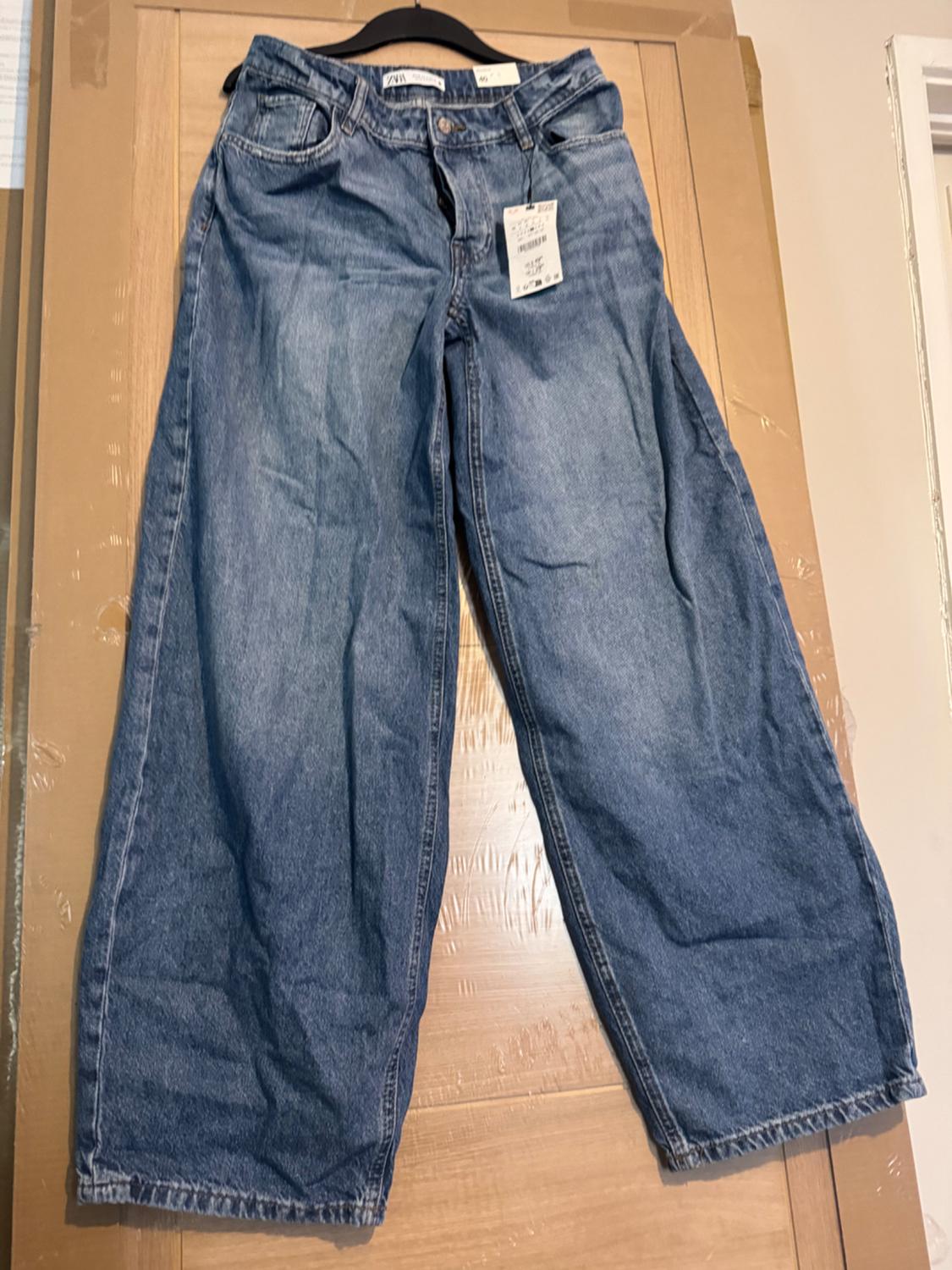 Blue wide-leg jeans hanging on a black hanger on a wooden door. Visible text: ZARA.