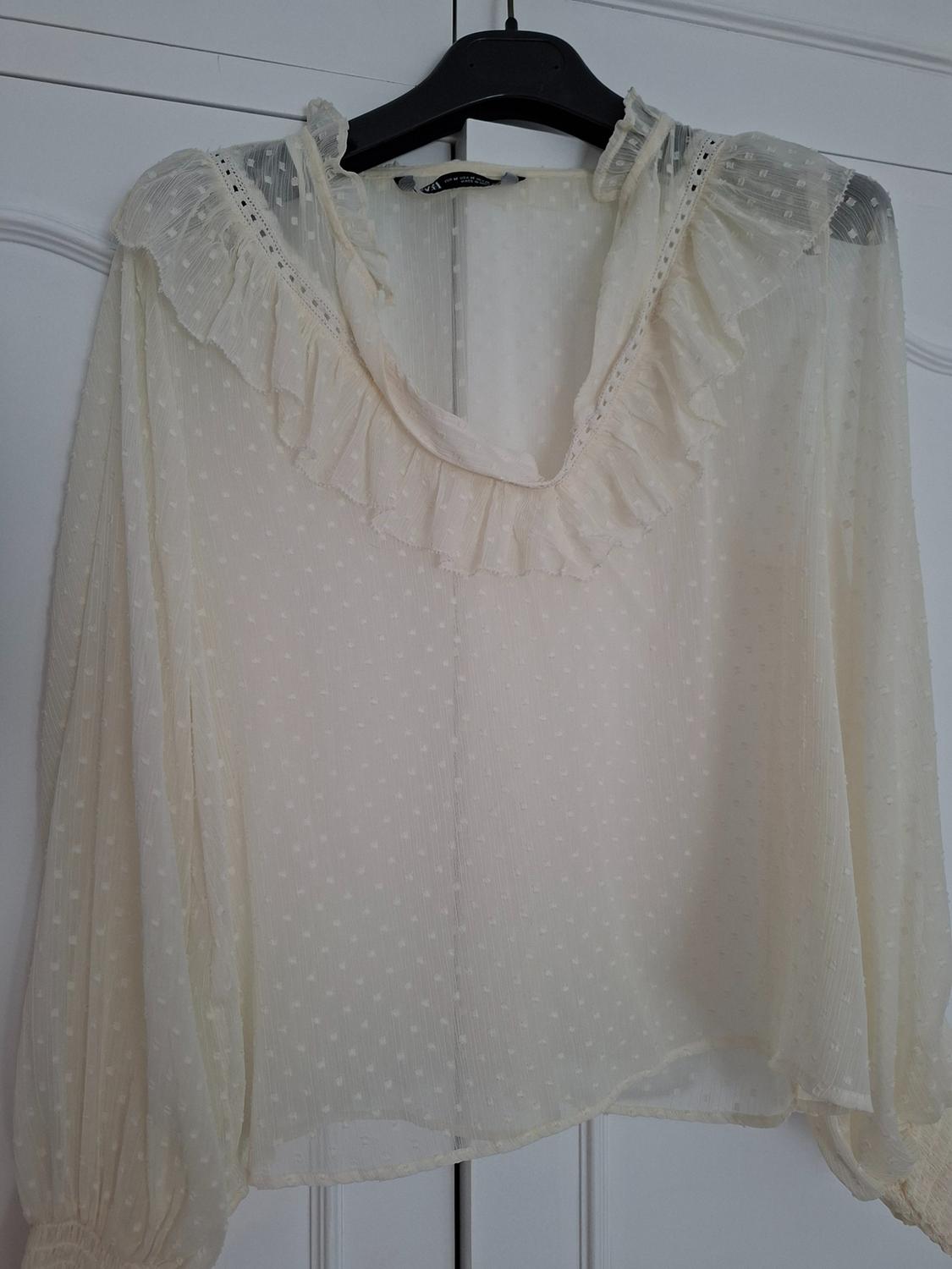 Cream semi-sheer blouse with a polka dot texture and a ruffled collar, hanging on a black hanger in front of a white wardrobe.