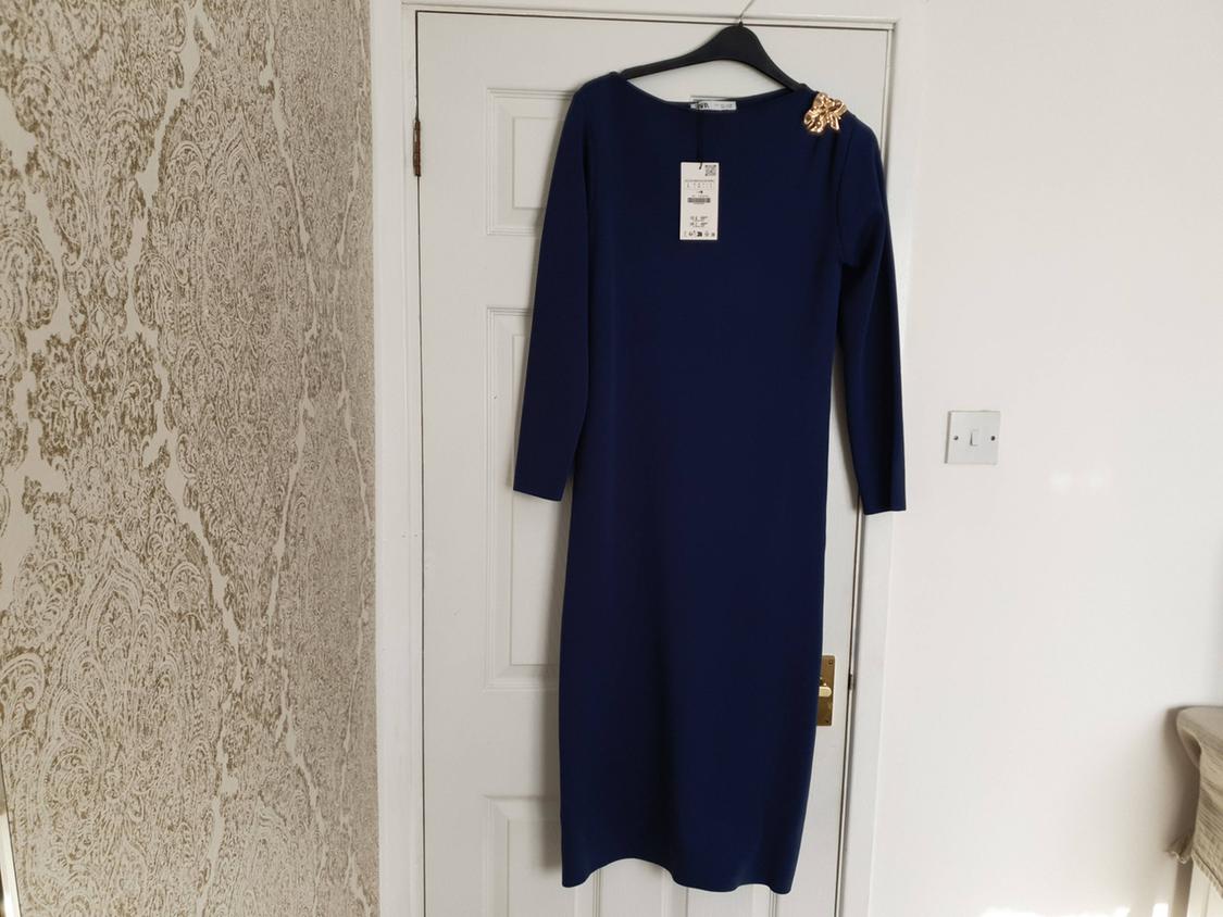 Navy blue long-sleeved dress with gold detail on the shoulder, hanging on a white door next to a wallpapered wall.