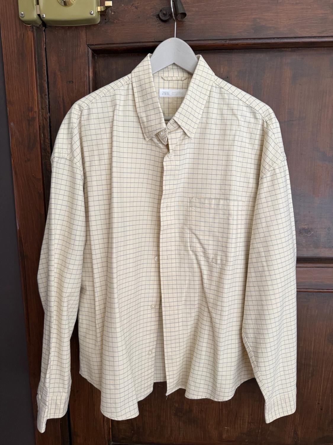 Beige checked shirt with long sleeves and a pocket, hanging on a hanger over a dark wooden door. Label with ZARA text.