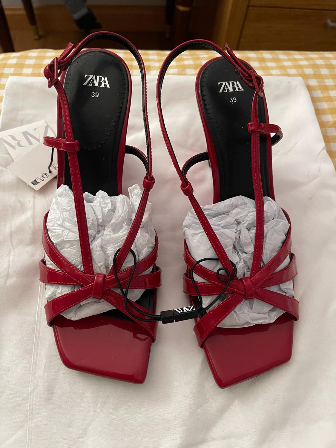 Pair of red strappy heeled sandals with a square toe on white fabric and a chequered background. Visible text: ZARA 39.