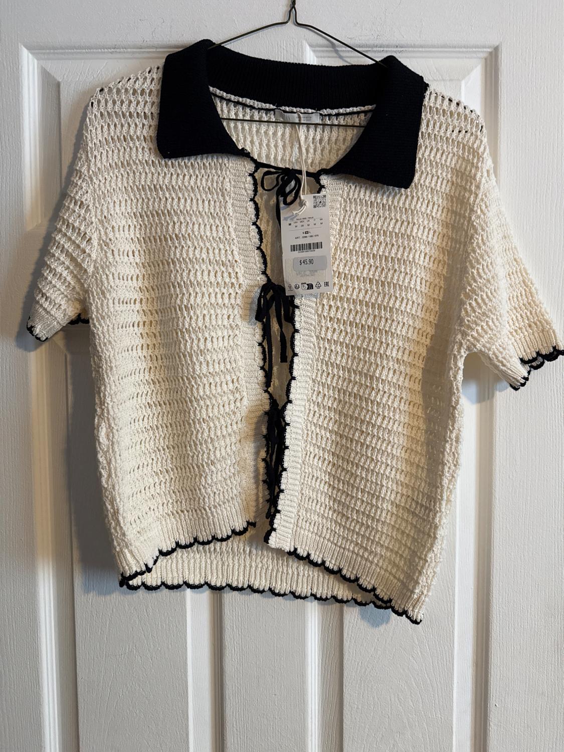 White knitted cardigan with black trim and contrasting collar, hanging on a hanger. Visible tag with price of $45.90.