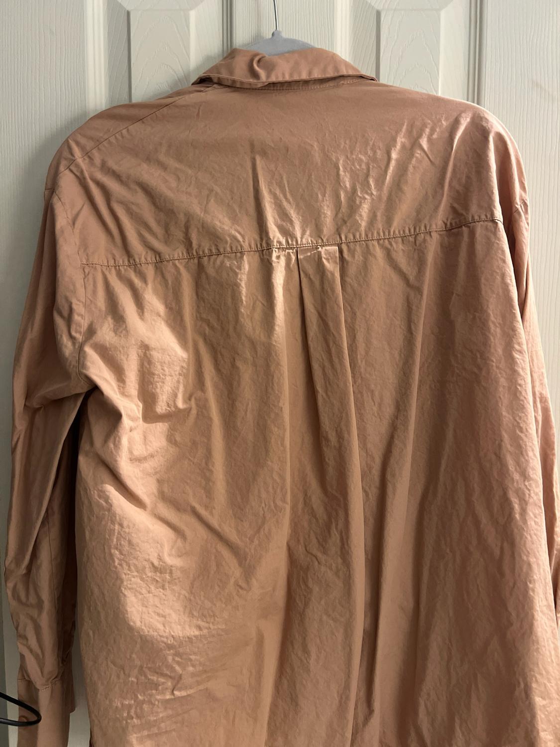 Back view of a salmon-colored shirt hanging on a hanger in front of a white door. The yoke and a central pleat in the back stand out.