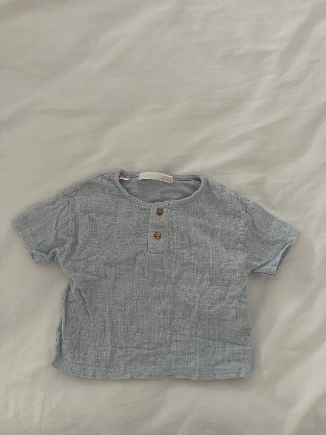 Light grey short-sleeved T-shirt with two wooden buttons at the neck, on a crumpled white surface.
