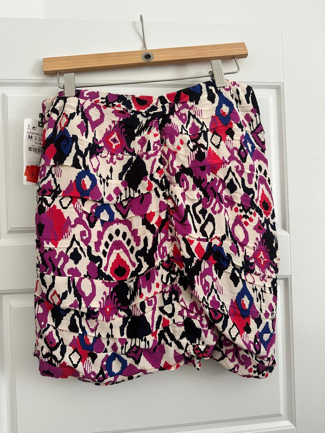 Short skirt with abstract print in shades of fuchsia, black, blue and white, hanging on a wooden hanger in front of a white door.