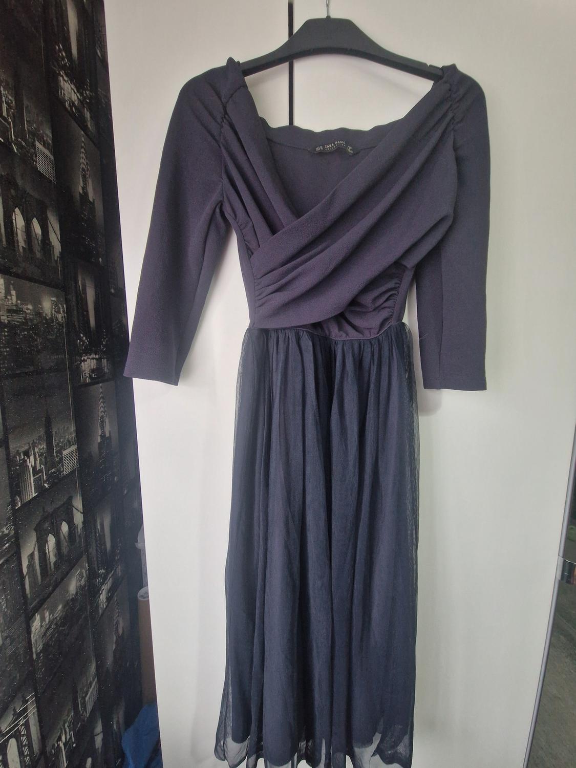 Black, long-sleeved dress hanging on a hanger, with a wrap neckline and a tulle skirt. Wall background with black and white photos.
