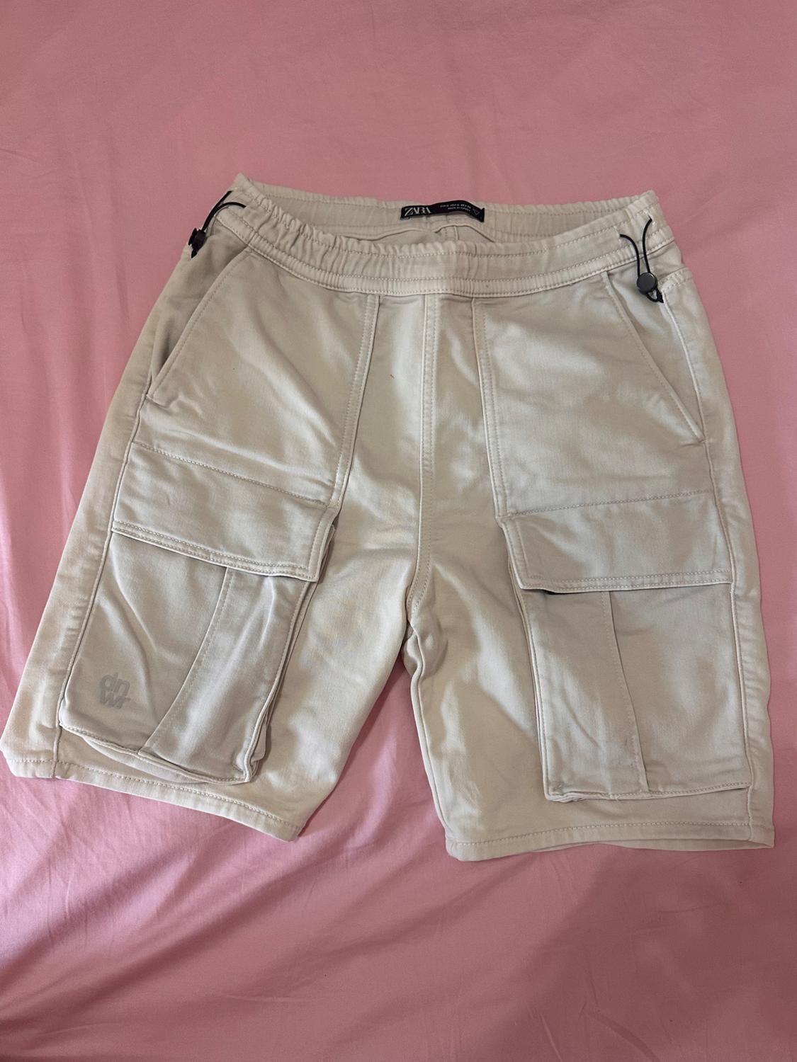 Beige shorts with large pockets on a pink surface. Elasticated waistband with black drawstrings. Visible label on the waistband.