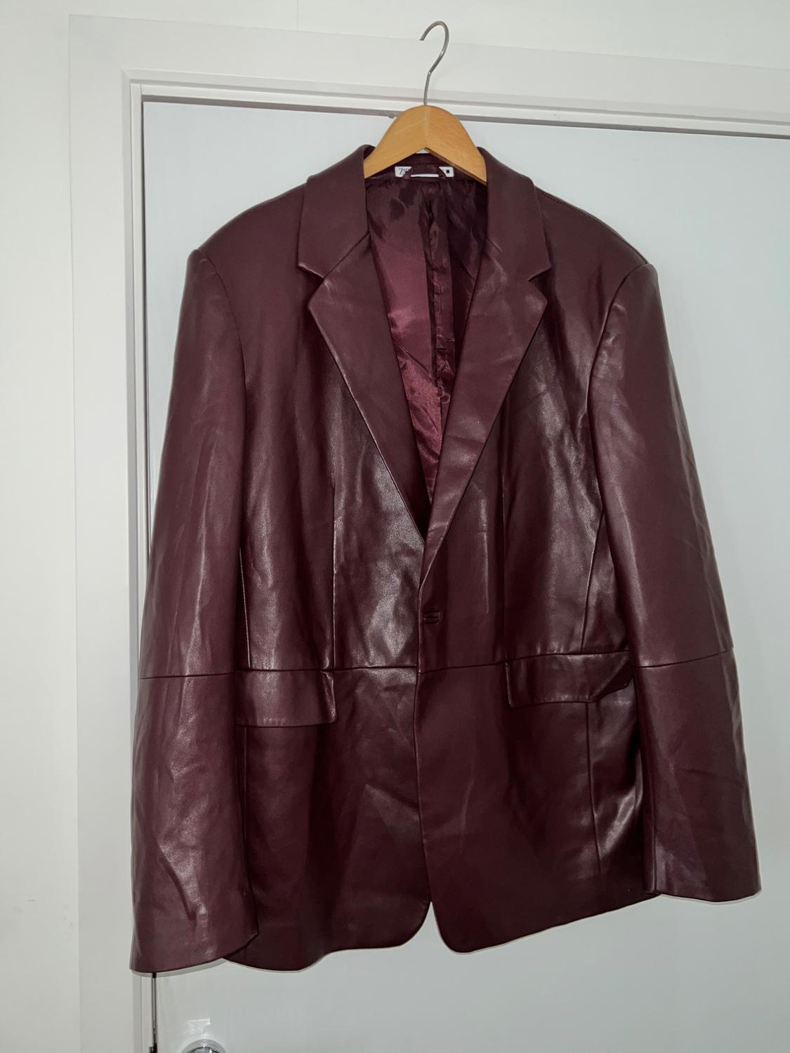Maroon faux leather blazer with flap pockets, hanging on a wooden hanger on a white door.