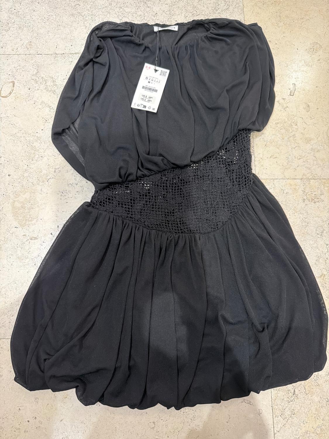Short black dress on a light-tiled floor. Draped bodice, crochet waist, and puffball skirt. Label with the letter S visible.