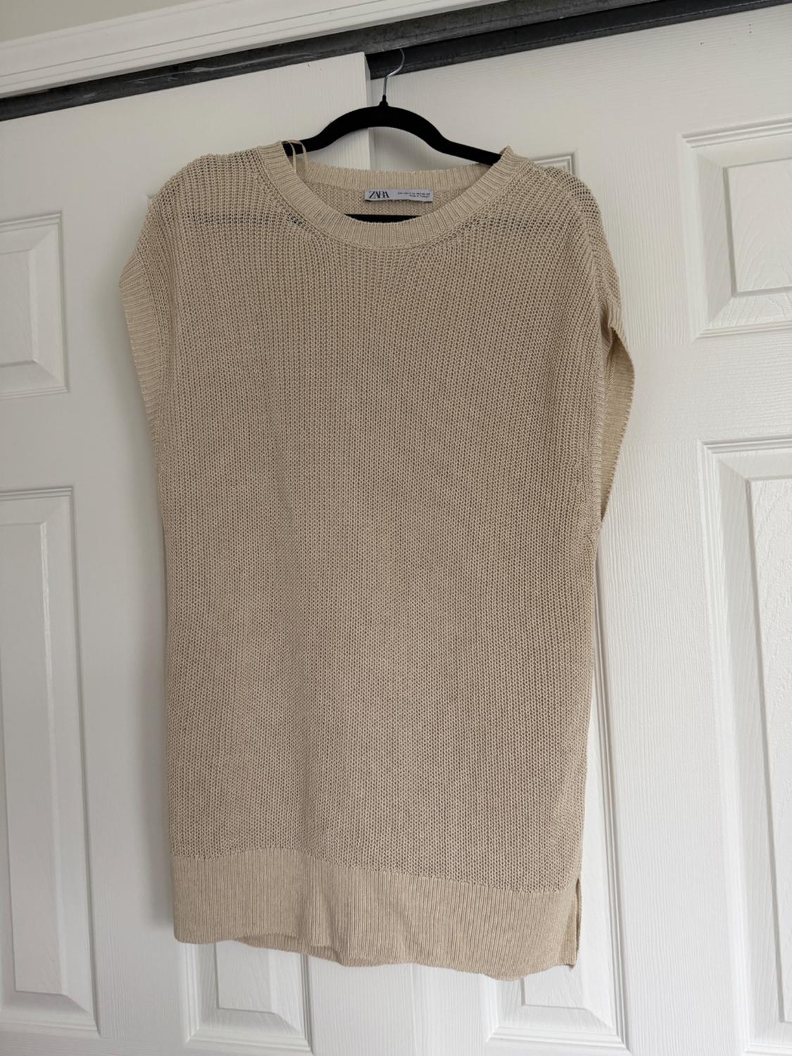 Beige knitted jumper hanging on a black hanger over a white door. Visible label with text "ZARA".