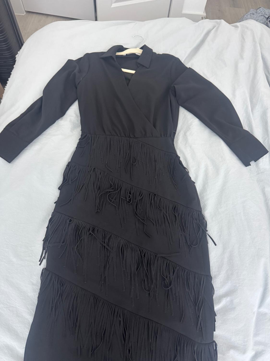 Black long-sleeved dress with a V-neck and a skirt with layers of fringing, laid on a bed with white sheets.