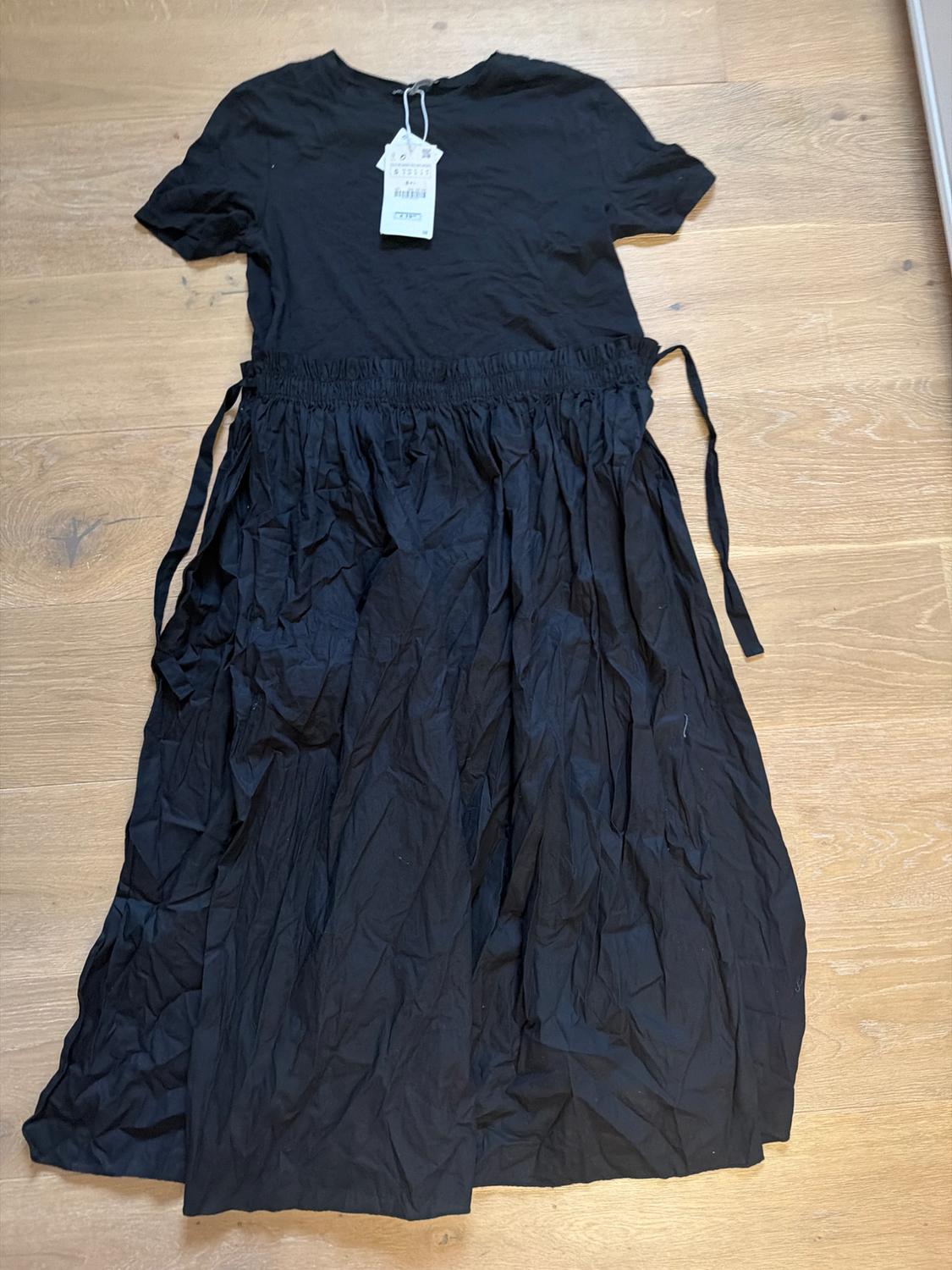 Black short-sleeved dress with a wrinkled pleated skirt on a wooden floor. Tags hanging from the collar.