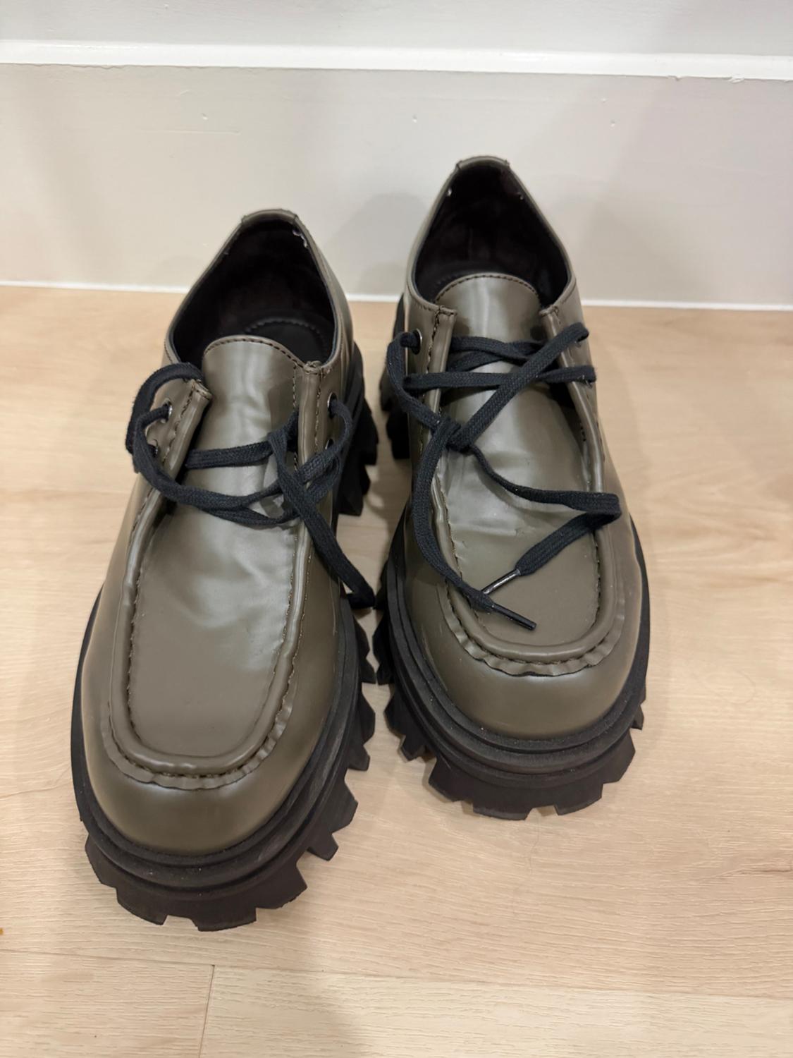 A pair of olive green shoes with a thick black sole and black laces, placed on a light wood floor in front of a white wall.