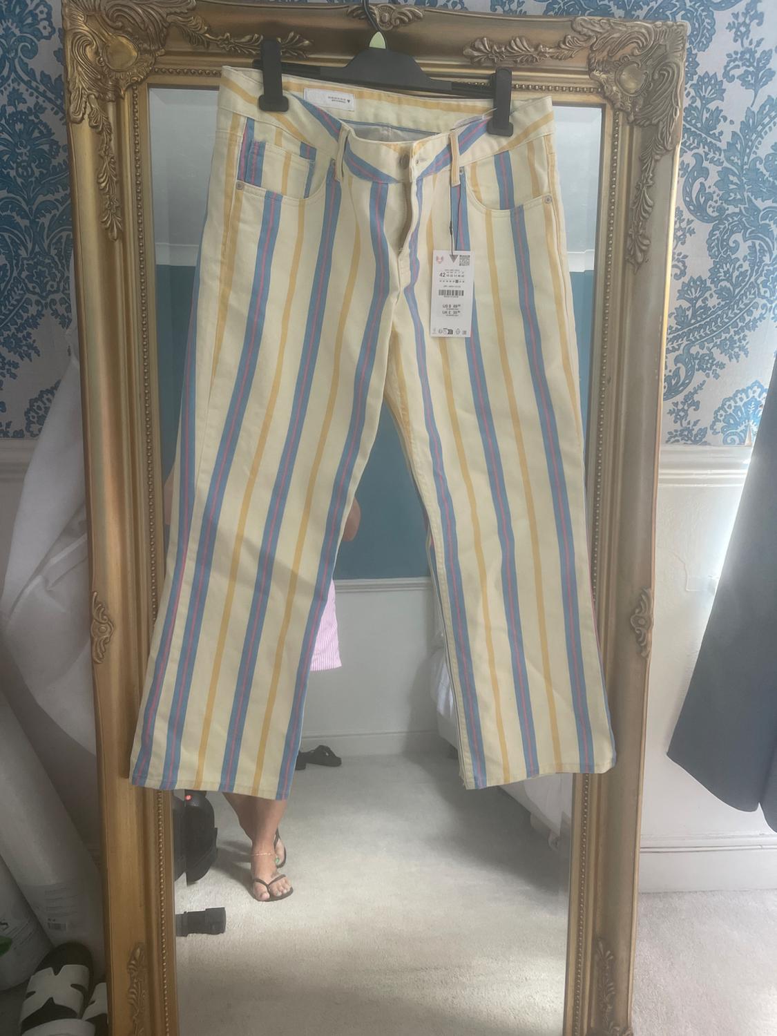 Yellow, blue and pink striped trousers hanging in front of a gold mirror that reflects a room and a person's legs.