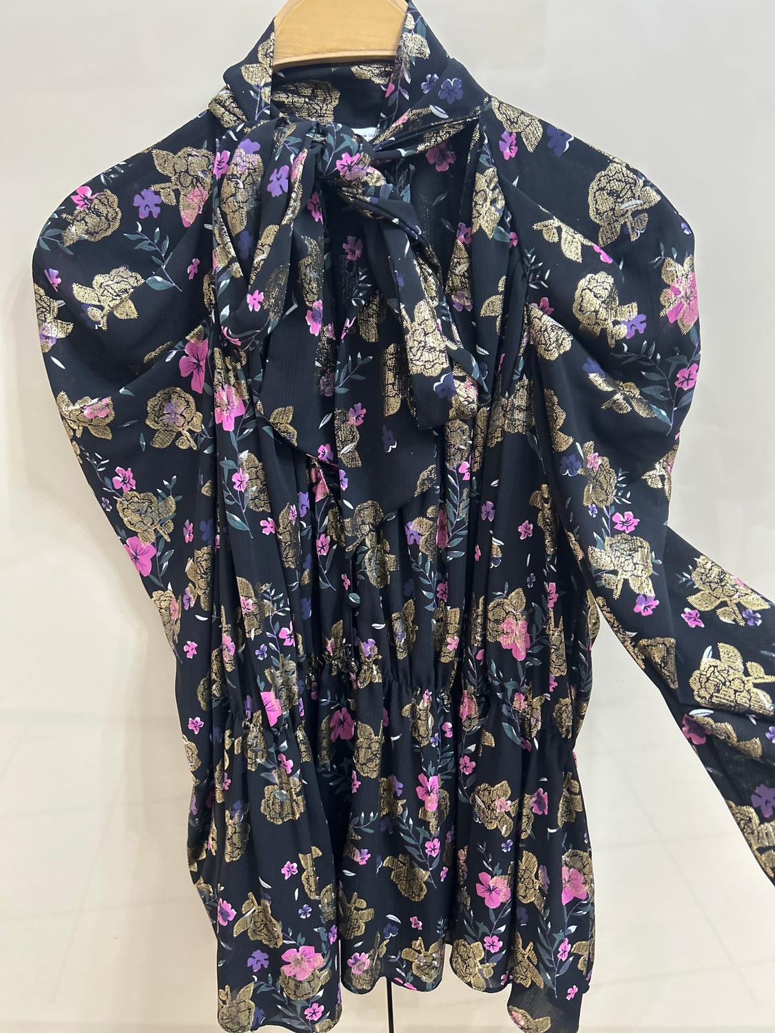 Long-sleeved blouse with a floral print in gold, pink, and purple tones on a black background, hanging on a wooden hanger.