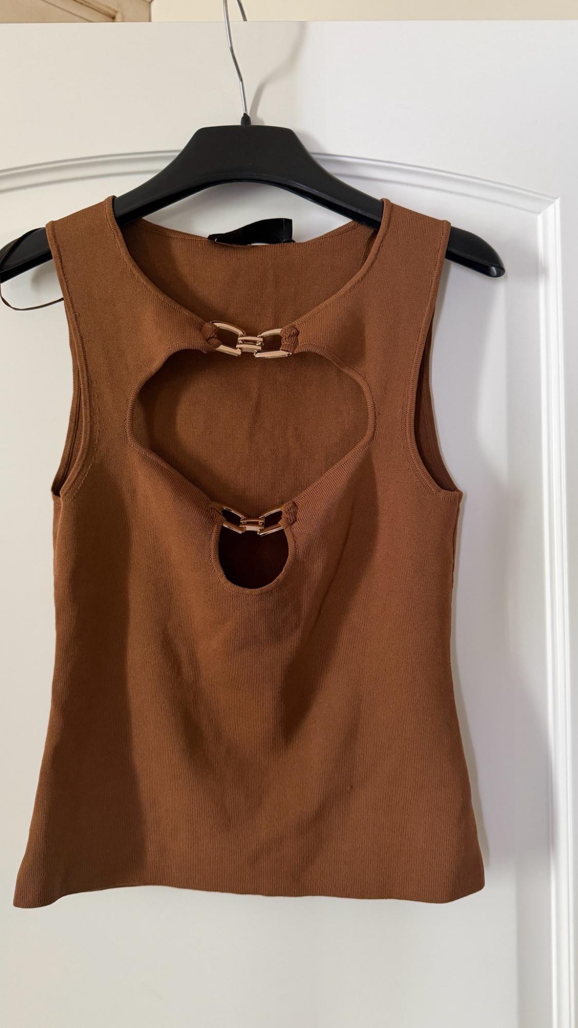 Brown sleeveless knitted top with cut-out details and gold-tone metal hardware, hanging on a black hanger in front of a white door.